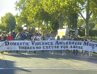 Photo of a group of people marching down a tree-lined street carrying a banner that says 'Domestic Violence Awareness Month: There's No Excuse for Abuse.'